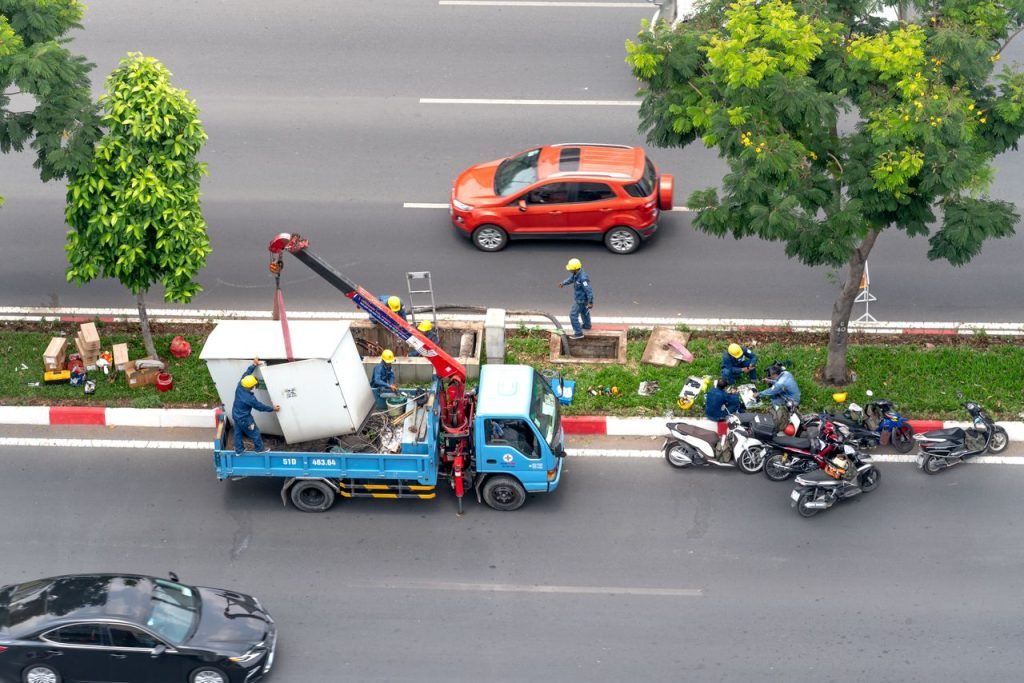 Workers perform maintenance on a busy road using a truck and crane, surrounded by motorcycles.